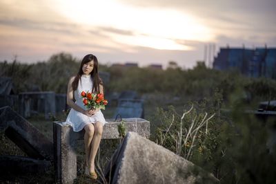 Portrait of young woman with flowers sitting on retaining wall against cloudy sky during sunset