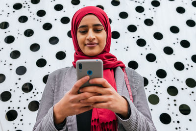 Side view of cheerful arab female in hijab standing sending message on mobile phone while smiling and communication on social media