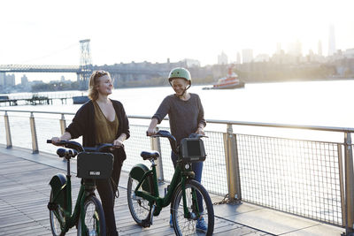 Smiling friends with bicycles walking on promenade by river