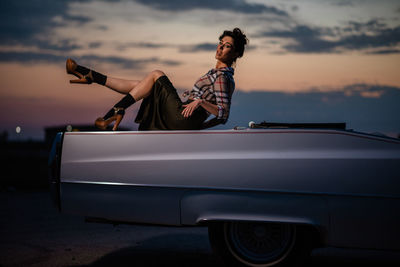 Portrait of woman standing by car against sky during sunset