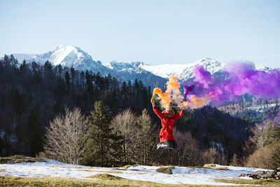 Full length of young woman jumping while holding distress flares against mountains