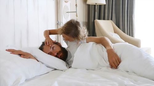 Cute girl opening eye of father lying on bed at home