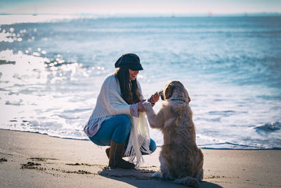 Full length of woman playing with dog while crouching on shore at beach