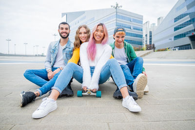 Group of teenagers fooling about at skate park - happy young friends sitting on skateboard 