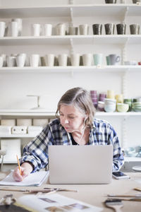 Mature female potter writing on paper while sitting with laptop at table in workshop