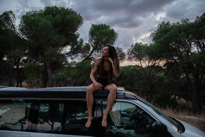 Woman sitting on car against trees
