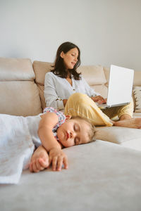 Little girl sleeping on the sofa while her mother works with a laptop and taking care of her