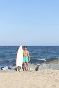 Shirtless young man with surfboard standing at beach against clear blue sky during sunny day