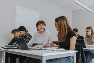 Smiling teenage girls sitting at desk by friends while studying together in classroom