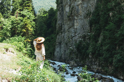 Young woman traveler with long blonde hair in straw hat looks at beautiful view of mountain river