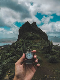 Cropped hand holding pocket watch near mountain against cloudy sky