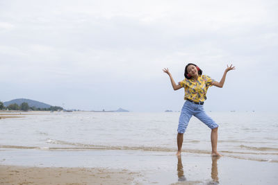 Girl dancing on music with headphones while standing on beach against sky