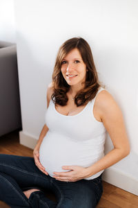 Portrait of smiling pregnant woman sitting against white wall at home