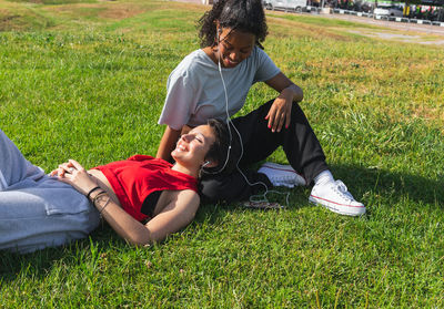 Cheerful multiracial young female friends in casual wear listening to music in earphones while resting on grassy lawn in park