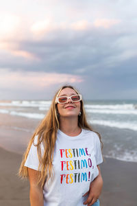 Positive young female in trendy sunglasses and stylish outfit standing at seaside against sea in summer evening