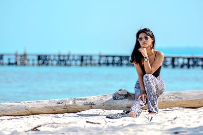 Full length of woman sitting on fallen tree trunk at beach against sky