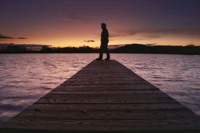 Silhouette woman standing on jetty by lake against sky during sunset