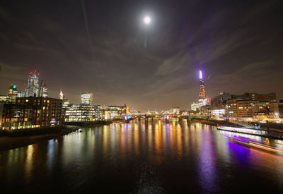 Illuminated buildings by river against sky at night