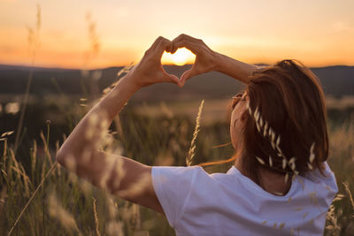 Rear view of woman making heart shape against sky during sunset