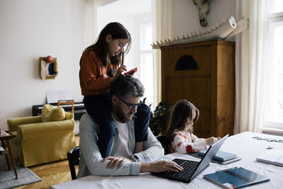 Daughter sitting on shoulder of father working on laptop at home