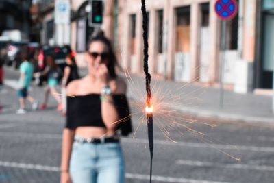 Young woman standing on city street with illuminated sparkler in foreground