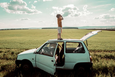 Playful boy standing on car roof with brother in vehicle at field against cloudy sky
