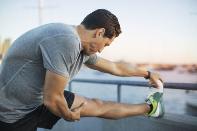Side view of determined athlete stretching leg on pier by harbor