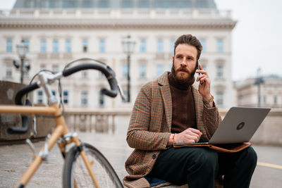 Businessman talking on phone while using laptop outdoors