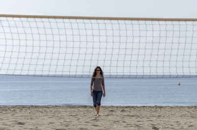 Woman walking at beach seen through volleyball net against sky