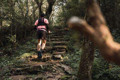 Back view of unrecognizable male traveler with trekking poles walking up old stone stairs in jungles