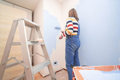 Standing woman dressed in overalls and striped blouse, seen from behind, painting a white wall