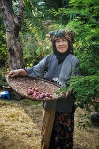 Portrait of senior woman holding onions in wicker basket against trees