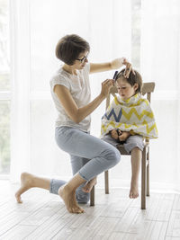 Mother cuts her son's hair by herself. little boy sits, covered with cloth, and holds  scissors. 