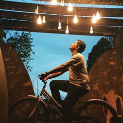 Side view of mature man looking at illuminated light bulbs while riding bicycle at sunset