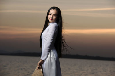 Beautiful woman standing at shore against sky during sunset