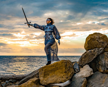 Full-length portrait of a woman medieval knight standing on a rock against a dramatic sky and a sea 