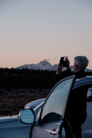 Woman photographing while standing by car against sky during sunset