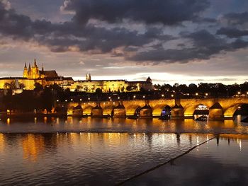 View of illuminated buildings by river against cloudy sky