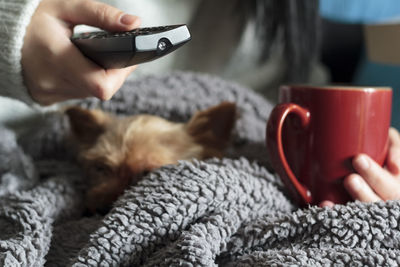Woman relaxing at home with her pet, holding hot drink and tv remote