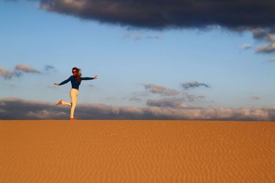 Woman standing on one leg with arms outstretched in desert against sky