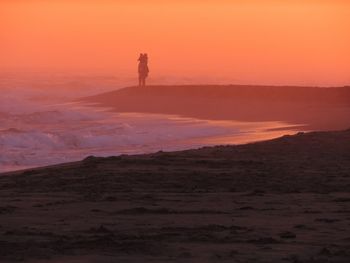 Silhouette person carrying kid while standing against sea during sunset