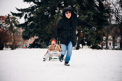 Caring father in sunglasses running while pulling happy kid on sledge while having fun on snowy street on winter day