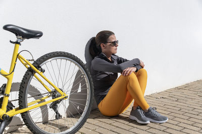 Woman sitting against a wall with her bicycle.