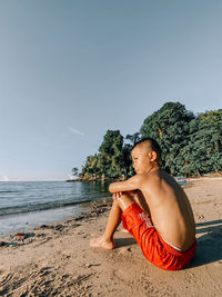 Young man sitting on shore at beach against sky