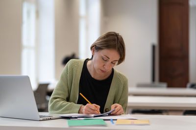 Middle-aged female student preparing for exam in public library while sitting at table with laptop