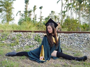 Young woman in graduation gown stretching on field by railroad track