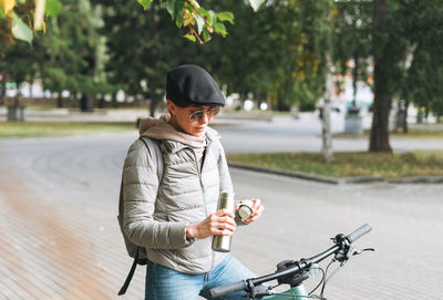 Portrait of woman in cap and sunglasses on bicycle with thermo mug on sunny autumn day in city