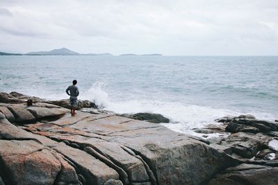 Rear view of man standing on rock formation by waves rushing against sky