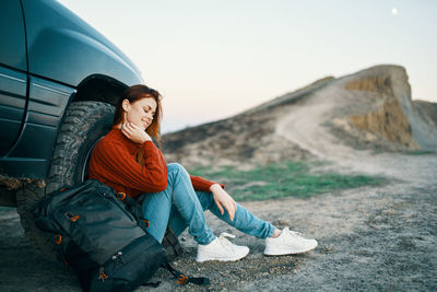 Full length of woman sitting by car against sky