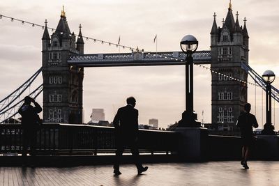 People walking on walkway against tower bridge during sunset